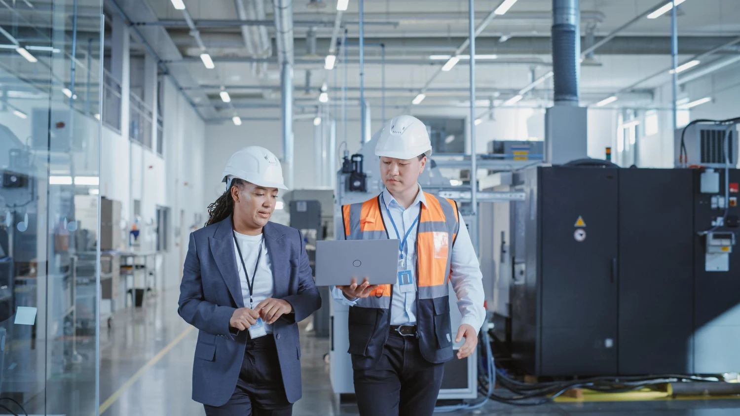 Profissionais com capacete analisando informações em laptop dentro de uma fábrica, representando gestão operacional e eficiência industrial.