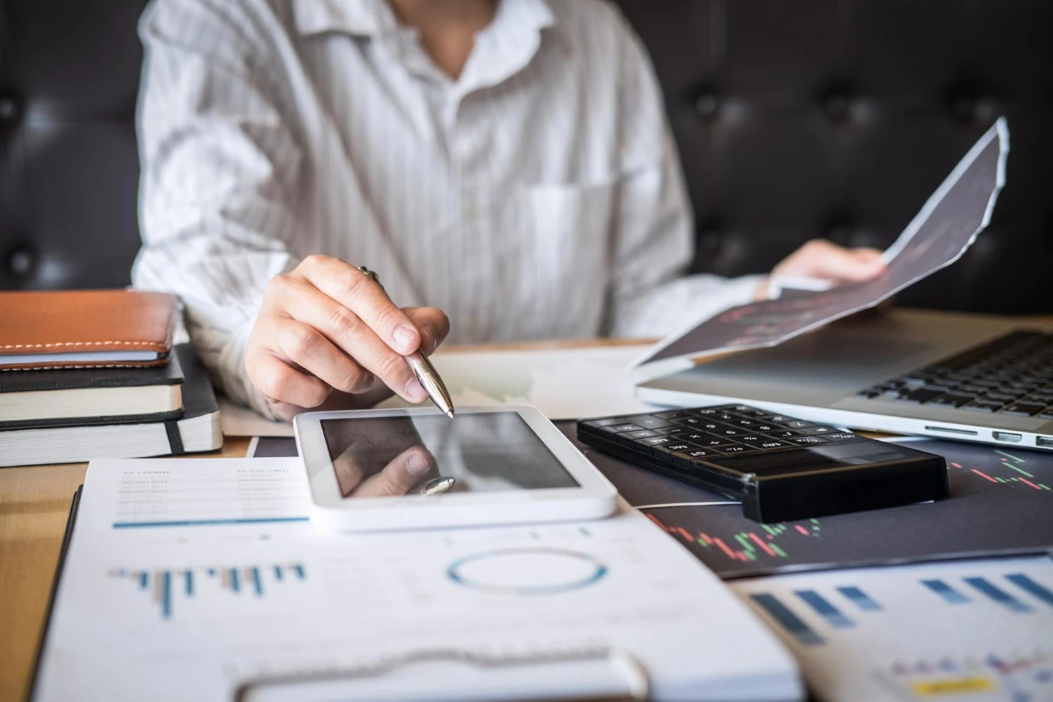 Pessoa analisando relatórios financeiros com tablet, documentos, calculadora e gráficos sobre a mesa.